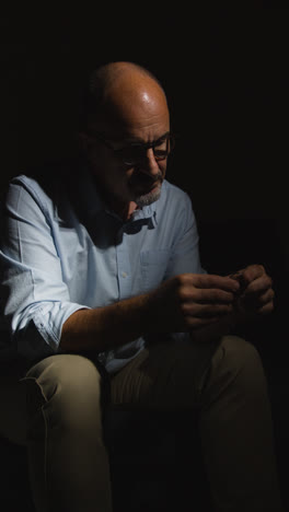 Vertical-Video-Of-Depressed-Divorced-Mature-Man-Sitting-In-Darkness-On-Sofa-Holding-Wedding-Ring-With-Low-Key-Lighting